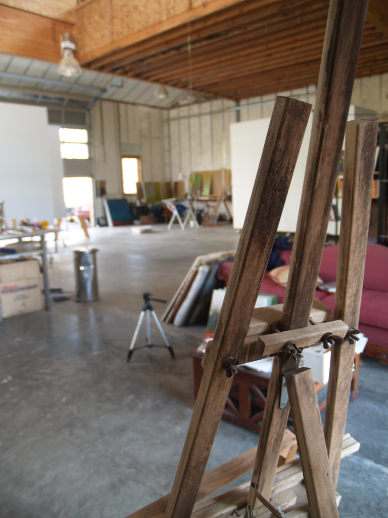 A working artist's studio interior: a wooden easel in warm ochre light, brushes and paint close at hand.